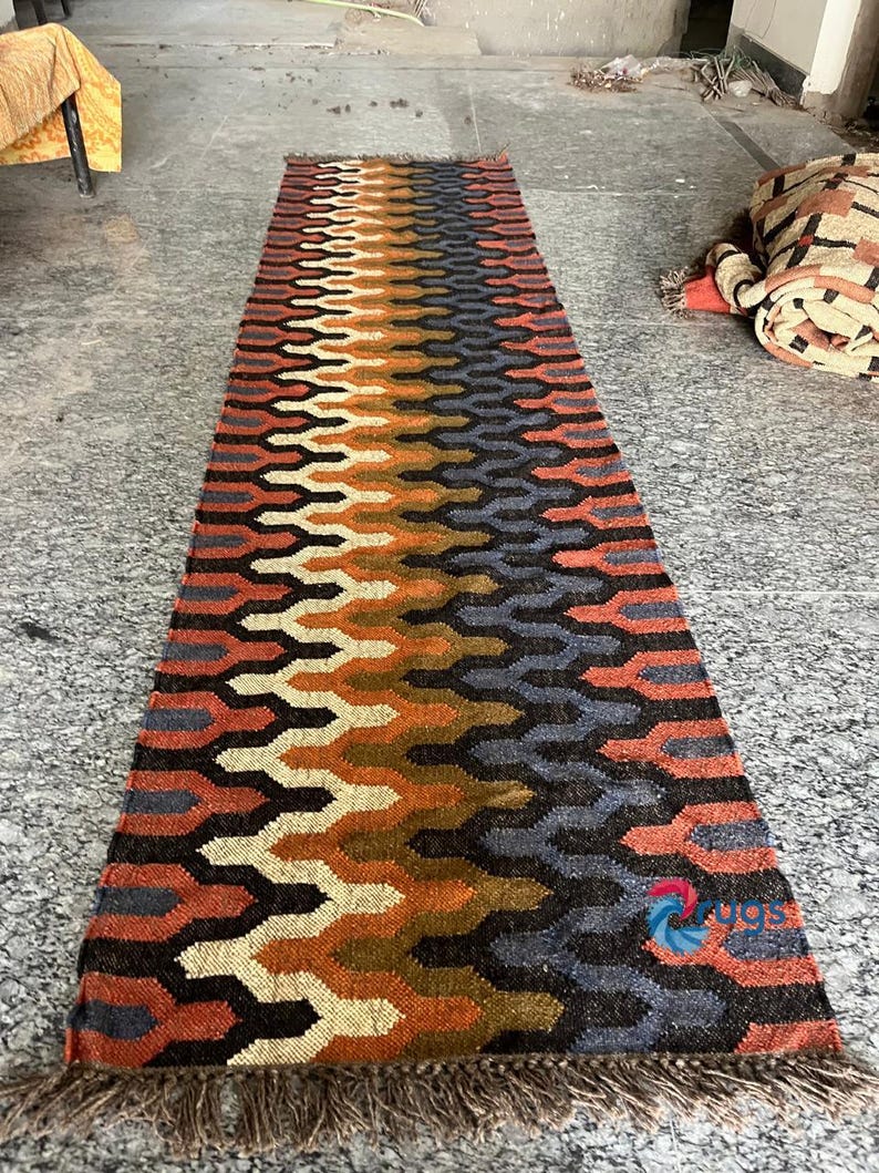 Puede incluir: Una alfombra rectangular con un patr&oacute;n geom&eacute;trico en tonos naranja, marr&oacute;n, negro y azul. La alfombra tiene un borde con flecos y un dise&ntilde;o repetitivo. La alfombra est&aacute; hecha de material tejido.