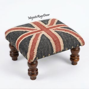 May include: A square footstool with a Union Jack design on the top. The fabric features a gray background with red and beige stripes. The wooden legs are dark brown and have a turned design. The text "Majestic Rugs Decor" is on the top.