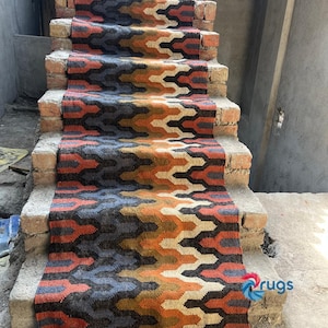 Puede incluir: Una colorida alfombra para escaleras con un patr&oacute;n geom&eacute;trico en tonos naranja, azul, marr&oacute;n y crema. La alfombra est&aacute; colocada sobre escalones de ladrillo y tiene un borde con flecos. Es un elemento decorativo para el hogar.