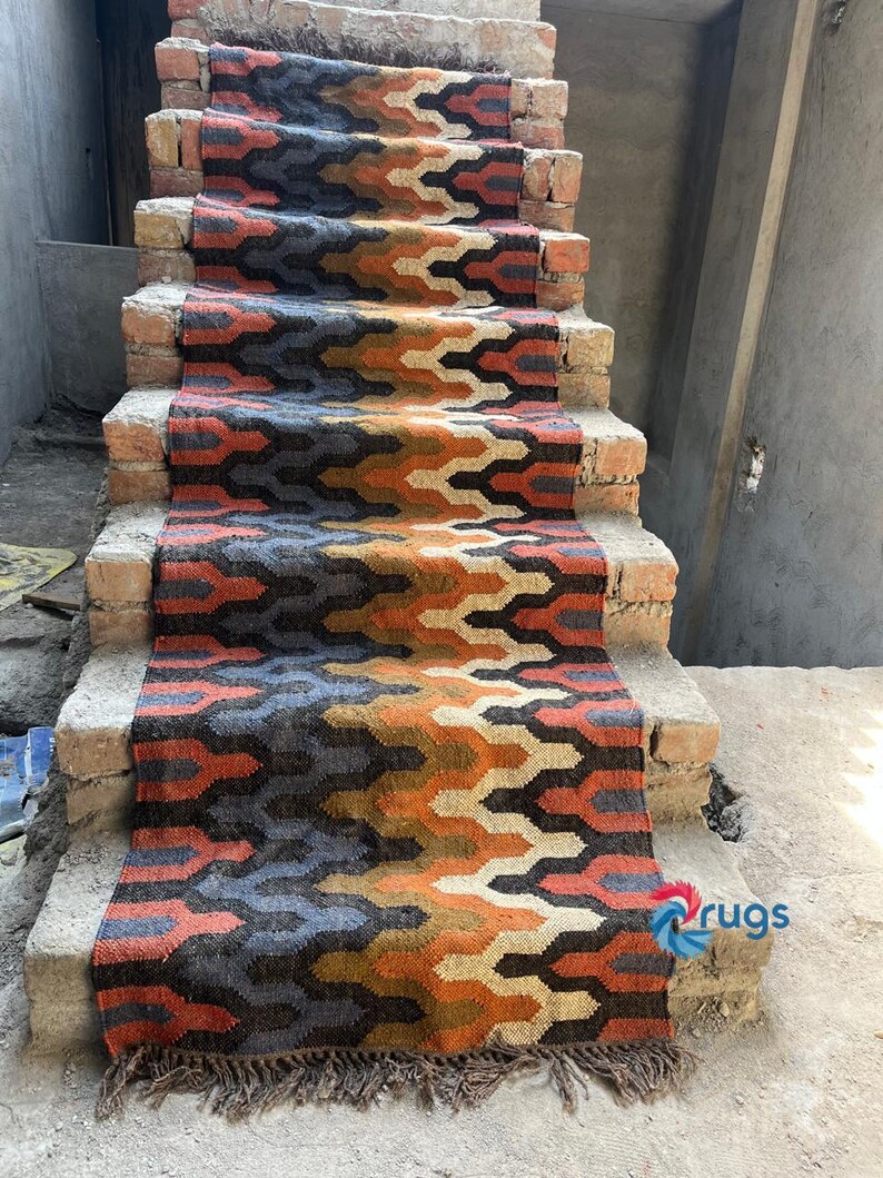 Puede incluir: Una colorida alfombra para escaleras con un patr&oacute;n geom&eacute;trico en tonos naranja, azul, marr&oacute;n y crema. La alfombra est&aacute; colocada sobre escalones de ladrillo y tiene un borde con flecos. Es un elemento decorativo para el hogar.