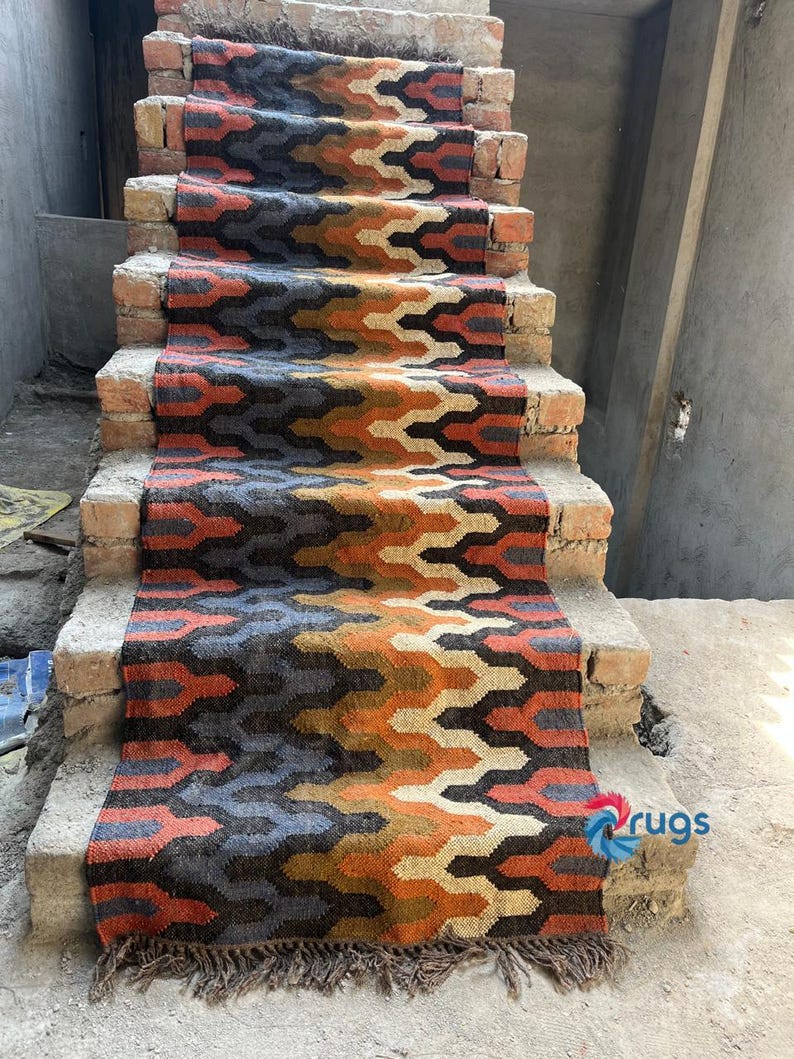 Puede incluir: Una colorida alfombra para escaleras con un patr&oacute;n geom&eacute;trico en tonos naranja, marr&oacute;n, gris y crema. La alfombra est&aacute; hecha de material tejido y tiene un borde con flecos. La alfombra se muestra en una escalera de ladrillo.