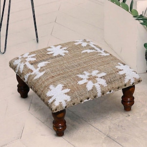 Op de afbeelding: Vierkante voetenbank met een bovenkant van natuurlijk geweven stof met witte bloemmotieven. De voetenbank heeft vier donkere houten poten en is afgewerkt met kleine metalen studs. Een decoratief stuk voor elke kamer.