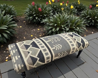 Puede incluir: Banco rectangular con una parte superior de tela gris, negra y beige estampada. El diseño incluye formas geométricas y motivos de flechas. El banco tiene cuatro patas torneadas negras. El texto "Majestic Rugs Decor" es visible.