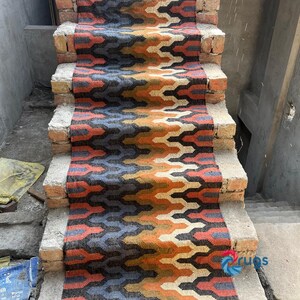 Puede incluir: Una colorida alfombra para escaleras con un patr&oacute;n geom&eacute;trico en tonos naranja, azul, negro y gris. La alfombra est&aacute; colocada sobre escalones de ladrillo, creando un contraste vibrante. El dise&ntilde;o presenta un motivo en zigzag repetitivo, agregando inter&eacute;s visual a la escalera.