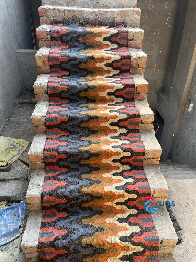 Puede incluir: Una colorida alfombra para escaleras con un patr&oacute;n geom&eacute;trico en tonos naranja, azul, negro y gris. La alfombra est&aacute; colocada sobre escalones de ladrillo, creando un contraste vibrante. El dise&ntilde;o presenta un motivo en zigzag repetitivo, agregando inter&eacute;s visual a la escalera.
