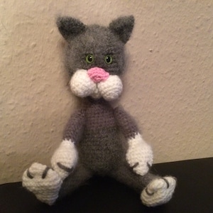 Könnte beinhalten: Ein graues Häkeltier in Form einer Katze mit weißen Pfoten und einer rosa Nase. Die Katze hat grüne Augen und sitzt auf einer dunklen Oberfläche.