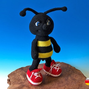 May include: A black and yellow crocheted bee wearing red sneakers. The bee has a friendly face with blue eyes and a yellow stripe across its abdomen.