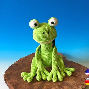 Könnte beinhalten: Ein grüner Häkel-Frosch sitzt auf einer Holzoberfläche. Der Frosch hat große weiße Augen mit schwarzen Pupillen und einen kleinen schwarzen Mund.