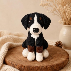 Op de afbeelding: Een gehaakt knuffeldier in de vorm van een hond met zwart, wit en bruin garen. De hond heeft een witte bles op het gezicht, zwarte oren, bruine vlekken boven de ogen en witte pootjes. Het heeft een zwarte plastic neus en donkere ogen.