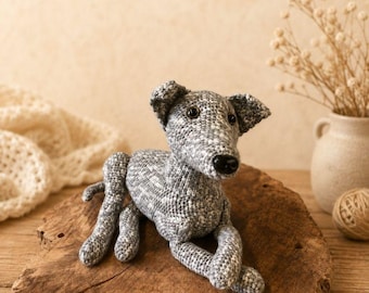 Puede incluir: Una figura de perro tejida a ganchillo a mano con hilo gris y blanco jaspeado. El peluche presenta un cuerpo esbelto, orejas caídas, un hocico puntiagudo con nariz de plástico negra y ojos de plástico marrón oscuro, en posición de reposo.