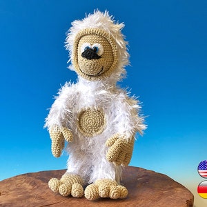 Op de afbeelding: Handgemaakte gehaakte Yeti-pop met een harige witte body en een bruin gezicht, handen en voeten. De Yeti heeft een lachende uitdrukking met blauwe ogen en een zwarte neus. De pop staat op een houten oppervlak tegen een blauwe achtergrond.