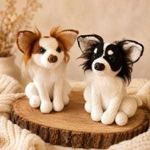 May include: Two crocheted dog figurines sitting side by side. The left dog is white with brown patches and long brown ear fur. The right dog is white with black patches and long black ear fur. Both have dark eyes and brown noses.