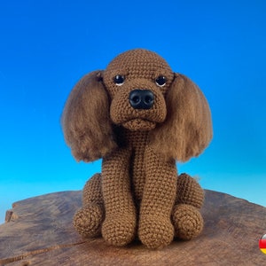 Könnte beinhalten: Ein handgehäkelter brauner Hund mit Schlappohren, einer schwarzen Nase und einem lieben Ausdruck. Der Hund sitzt auf einer Holzoberfläche vor blauem Hintergrund. Der Hund ist ein Cocker Spaniel.