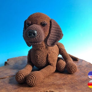 Puede incluir: Un juguete de perro de crochet marrón sentado en una superficie de madera. El perro tiene ojos marrones y una nariz negra.
