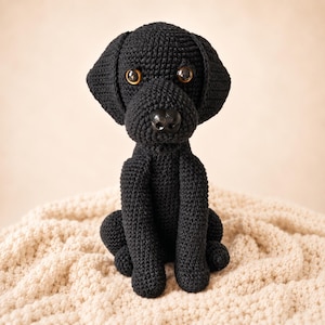 Könnte beinhalten: Eine schwarze Hundefigur aus gehäkeltem Garn. Der Hund hat eine texturierte, gestickte Oberfläche, zwei große braune Augen und eine glatte schwarze Kunststoffnase. Er ist in sitzender Position mit nach vorne gerichteten Vorderpfoten dargestellt.