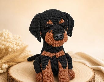 May include: A handcrafted crochet dog figurine featuring a black and tan color scheme. The plush toy has floppy ears, a rounded snout with a black plastic nose, and dark eyes. The yarn texture is visible throughout the seated canine figure.
