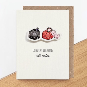 Könnte beinhalten: Eine weiße Grußkarte mit einem schwarzen zwanzigseitigen Würfel mit Zylinder und einem roten zwanzigseitigen Würfel mit Blumenkranz und Schleier. Der Text CONGRATULATIONS roll mates! ist in schwarzer Schrift unter den beiden Würfeln abgedruckt.