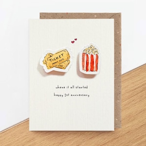 May include: A white greeting card featuring a watercolour illustration of two yellow movie tickets and a red and white striped popcorn bucket. Two small red hearts are above the items. The text reads: where it all started happy 1st anniversary.