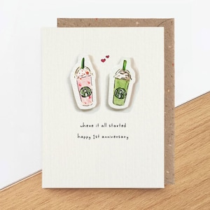 May include: A white greeting card featuring two illustrated beverage cups, one pink and one green, with two small red hearts above them. The text reads: where it all started, happy 1st anniversary. The card has a textured surface and a brown envelope.