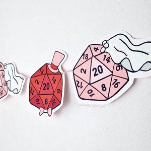 May include: A garland of illustrated D20 dice, some adorned with wedding attire. The dice are in shades of pink and red, with numbers visible on each face. The garland is strung on a pink string, perfect for a wedding or gaming-themed event.