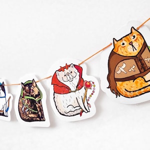 May include: A decorative garland featuring illustrated cats in costumes, strung on orange twine. The cats are dressed as a devil, a ranger, and an archer. The illustrations are colourful and whimsical, set against a white background.