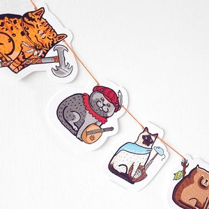 Könnte beinhalten: Eine verspielte Girlande mit illustrierten Katzenfiguren in verschiedenen Kostümen. Die Katzen sind mit einer Axt, einer Laute und einer Maus dargestellt. Die Girlande ist auf einer dünnen orangefarbenen Schnur aufgereiht, ideal zum Dekorieren.