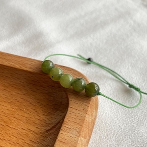 May include: A green beaded bracelet with five round, olive-green beads strung on a thin, green cord. The bracelet is displayed on a wooden tray with a white cloth background. The beads are approximately 0.5 inches in diameter.