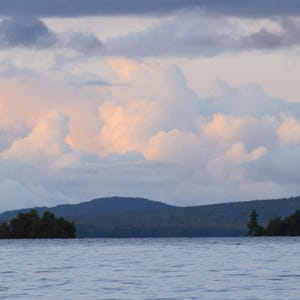 Puede incluir: Una vista panorámica de un lago con una costa arbolada y un cielo nublado. Las nubes son de un suave azul claro y rosa, creando una atmósfera pacífica y serena.