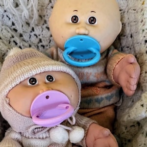 May include: Two dolls with round faces and brown eyes. One doll is bald and wears a tan sweater with a blue pacifier. The other doll wears a tan knit hat with two white pom-poms, a matching tan sweater, and a light purple pacifier in its mouth.