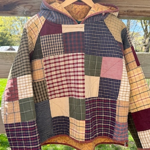 May include: A patchwork hooded jacket in a variety of plaid and patterned fabrics, including red, blue, green, and tan. The jacket is quilted and has a hood with a contrasting lining. It is hanging on a wooden hanger.