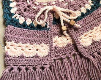 Op de afbeelding: Een handgemaakt gehaakt vest in tinten mauve, blauwgroen en crème. Het vest heeft een gefranjerde zoom, geschulpte randen en een trekkoordsluiting met houten kralen. Het ingewikkelde haakwerk creëert een textuurrijk en gedetailleerd ontwerp.