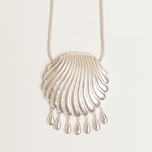 Puede incluir: Collar con colgante plateado con un diseño de concha texturizada. El colgante tiene una serie de colgantes en forma de lágrima. La cadena del collar es una fina cadena de serpiente plateada.