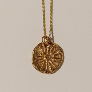 May include: A gold-toned necklace featuring a circular pendant with a textured, irregular surface and a raised sunburst or starburst pattern radiating from the center. The pendant hangs from a delicate, thin gold-toned chain with small links.