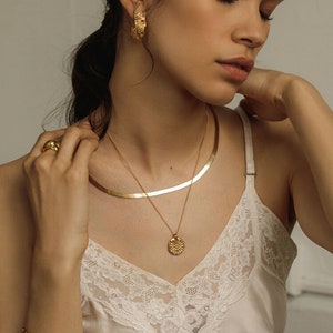 May include: A gold-toned jewelry set consisting of a flat herringbone chain necklace, a delicate chain necklace with a round sunburst pendant, textured hoop earrings, a smooth band ring, and a rectangular link bracelet worn with a white lace camisole.