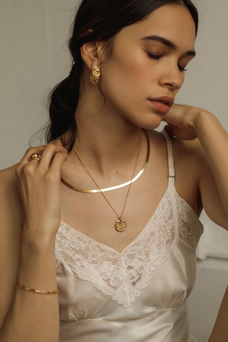 May include: A gold-toned jewelry set consisting of a flat herringbone chain necklace, a delicate chain necklace with a round sunburst pendant, textured hoop earrings, a smooth band ring, and a rectangular link bracelet worn with a white lace camisole.