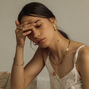 May include: A set of gold-toned jewelry including a textured hoop earring, a flat herringbone chain necklace, a delicate chain necklace with a round pendant, a link bracelet, and a ring with a textured surface, worn with a white lace-trimmed camisole.