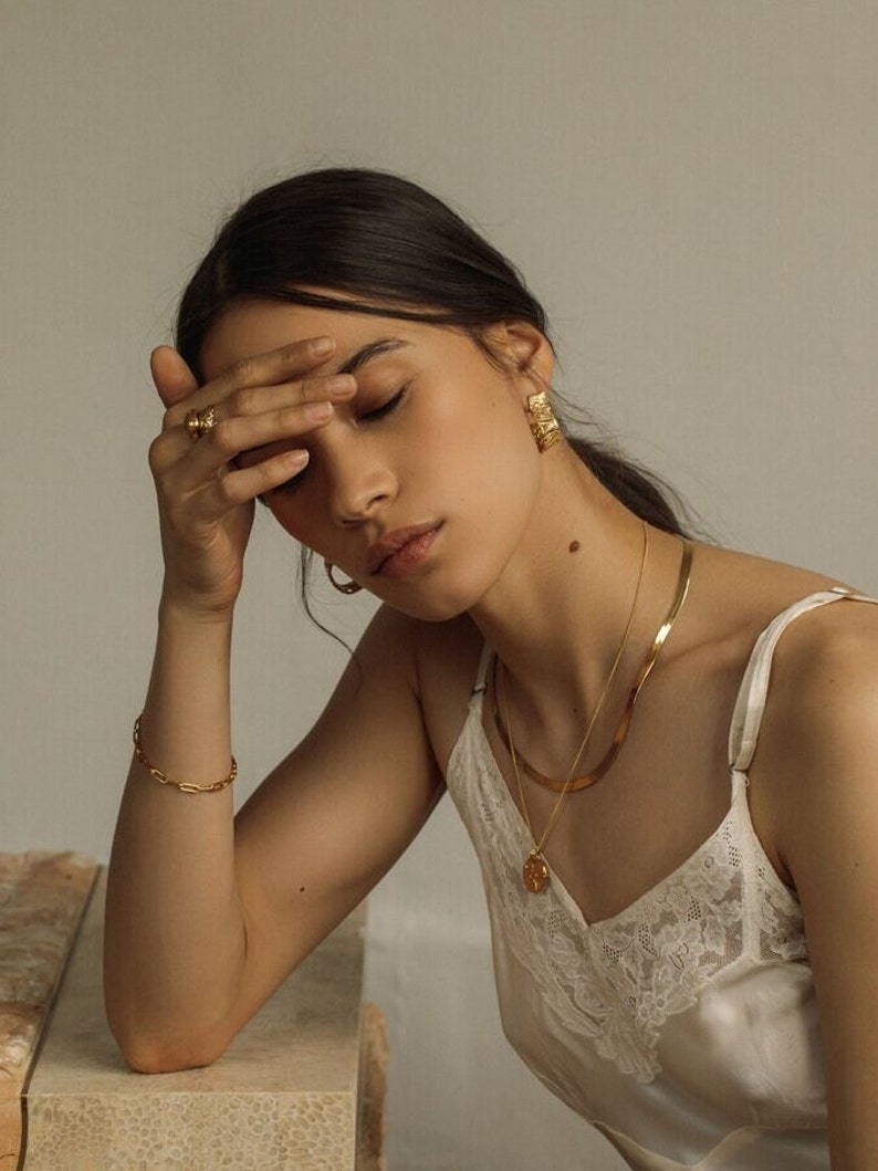 May include: A set of gold-toned jewelry including a textured hoop earring, a flat herringbone chain necklace, a delicate chain necklace with a round pendant, a link bracelet, and a ring with a textured surface, worn with a white lace-trimmed camisole.