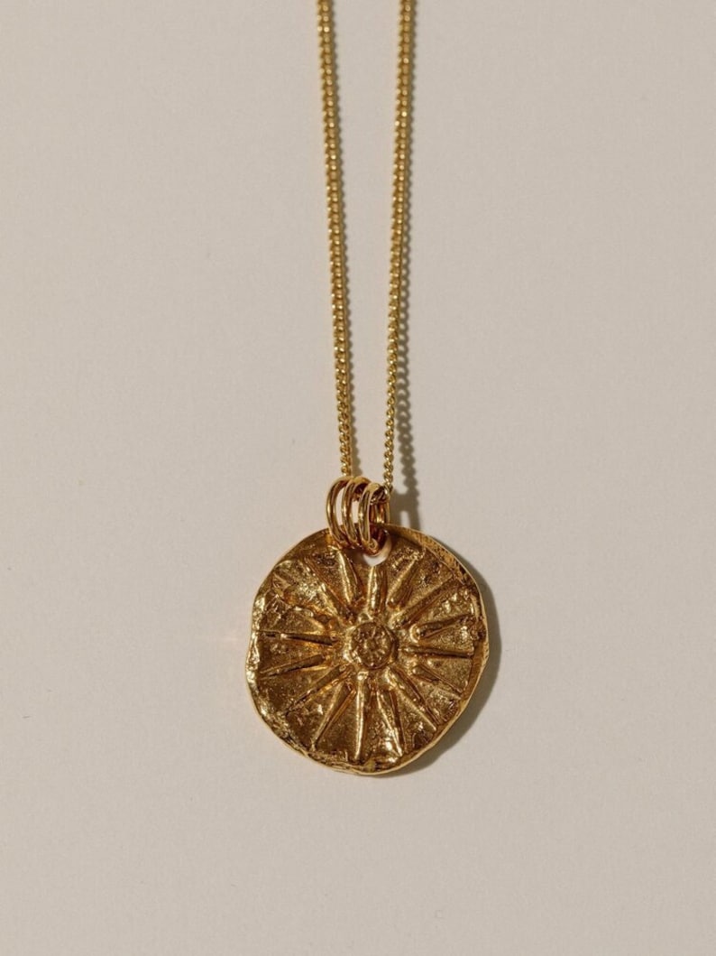 May include: A gold-toned necklace featuring a circular pendant with a textured, irregular surface and a raised sunburst or starburst pattern radiating from the center. The pendant hangs from a delicate, thin gold-toned chain with small links.