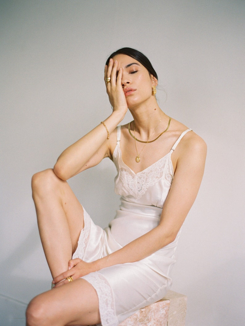May include: A cream-colored satin slip dress featuring delicate white lace trim along the V-neckline and hem. The garment has thin spaghetti straps and a smooth, lustrous finish. Gold jewelry including a necklace, bracelet, and rings are worn.
