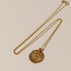 May include: A gold-toned necklace featuring a thin, delicate chain and a circular pendant. The pendant has a textured, uneven surface with a raised sunburst or star-like design radiating from the center. The necklace includes a small circular clasp.