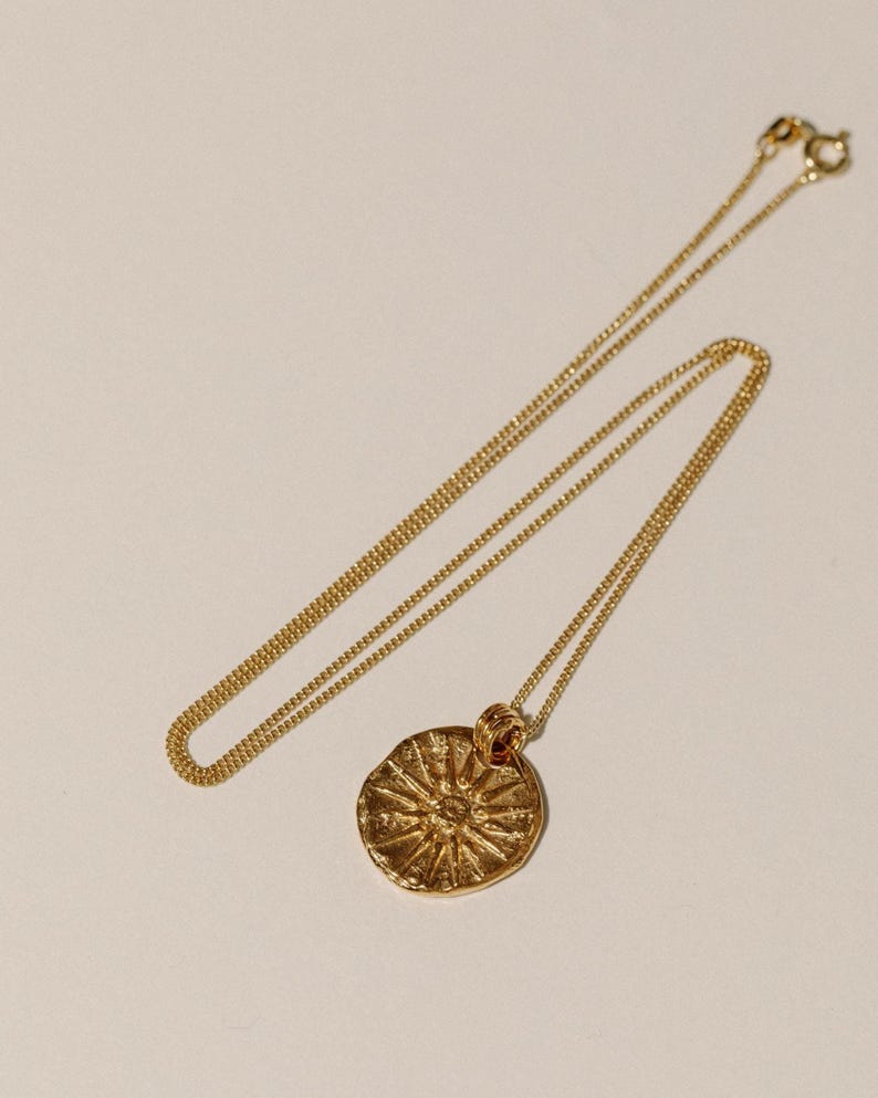 May include: A gold-toned necklace featuring a thin, delicate chain and a circular pendant. The pendant has a textured, uneven surface with a raised sunburst or star-like design radiating from the center. The necklace includes a small circular clasp.