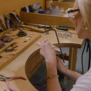 May include: A jeweler's workbench featuring a metal jeweler's saw, a wooden bench pin, various metal pliers with pink and black handles, a wooden mallet, and small metal components scattered across the wooden surface for jewelry fabrication.