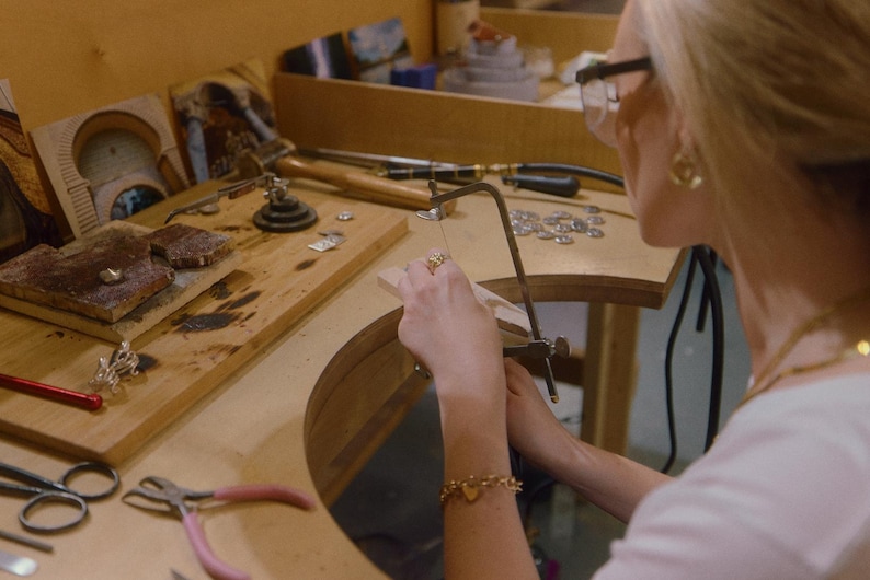 May include: A jeweler's workbench featuring a metal jeweler's saw, a wooden bench pin, various metal pliers with pink and black handles, a wooden mallet, and small metal components scattered across the wooden surface for jewelry fabrication.