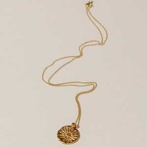 May include: A gold-toned necklace featuring a delicate chain and a circular pendant. The pendant has a textured, uneven surface with a raised sunburst or star-like pattern radiating from the center. The metal has a polished, reflective finish.