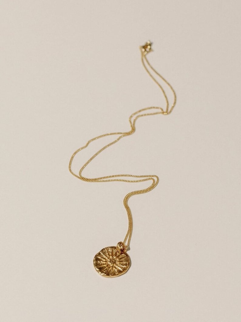 May include: A gold-toned necklace featuring a delicate chain and a circular pendant. The pendant has a textured, uneven surface with a raised sunburst or star-like pattern radiating from the center. The metal has a polished, reflective finish.