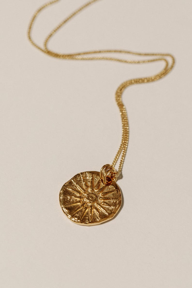 May include: A gold-toned necklace featuring a circular pendant with a textured, sunburst-like design radiating from the center. The pendant is suspended from a delicate, fine-link gold chain that loops through a small, coiled metal bail at the top.