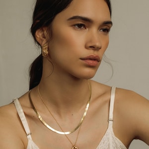 May include: A gold-toned jewelry set consisting of a flat herringbone chain necklace, a delicate chain necklace with a round textured pendant featuring a sunburst design, and a pair of matching textured gold-toned stud earrings.