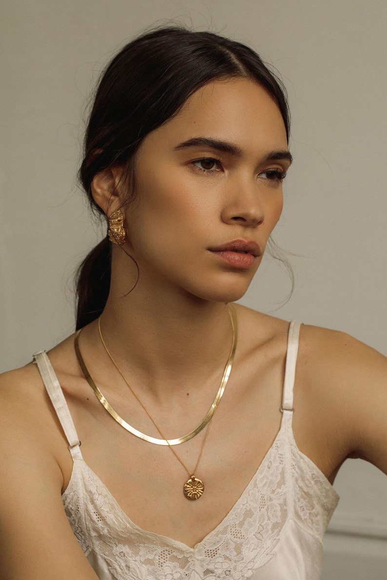 May include: A gold-toned jewelry set consisting of a flat herringbone chain necklace, a delicate chain necklace with a round textured pendant featuring a sunburst design, and a pair of matching textured gold-toned stud earrings.