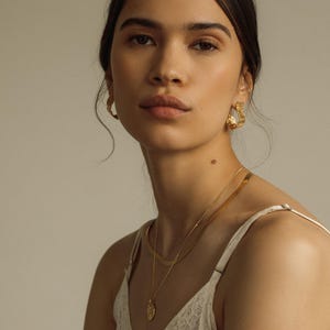 May include: A pair of gold hoop earrings with a textured, organic surface finish, paired with a layered gold necklace set consisting of a flat herringbone chain and a delicate chain featuring a small, round, textured gold pendant charm.