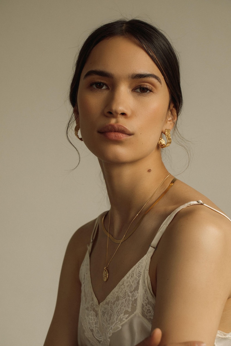 May include: A pair of gold hoop earrings with a textured, organic surface finish, paired with a layered gold necklace set consisting of a flat herringbone chain and a delicate chain featuring a small, round, textured gold pendant charm.
